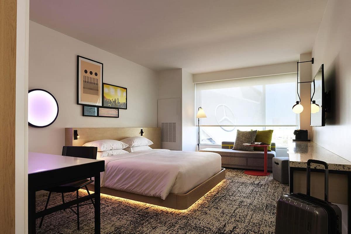 A modern hotel room with a large bed, a desk, a chair, a small table, a red stool, and a suitcase.