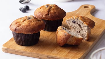 Three muffins on a wooden board, one cut in half, with a bowl of blueberries in the background.