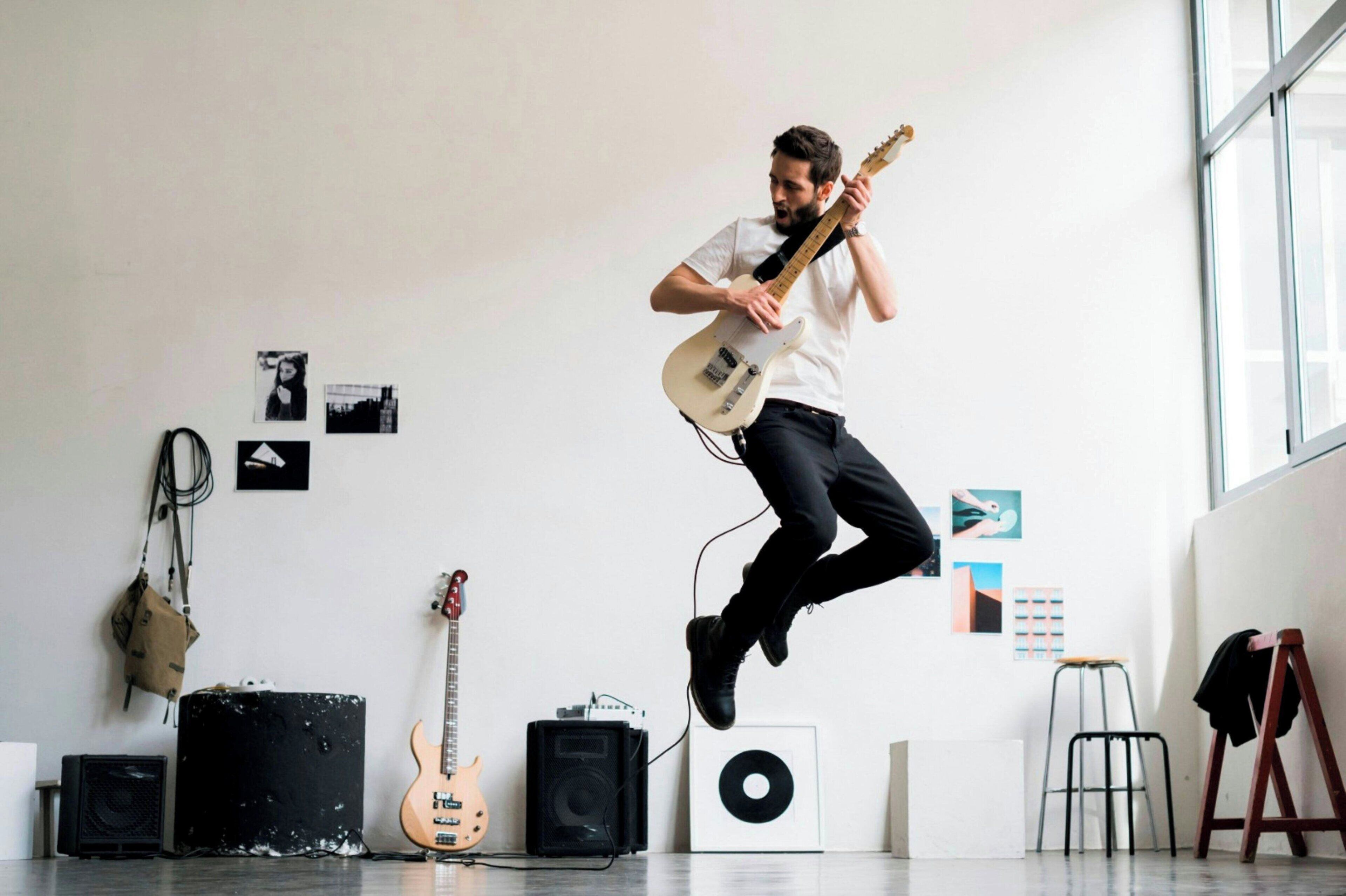 Una persona tocando una guitarra eléctrica en una habitación con pared blanca, una ventana y diverso equipo musical.