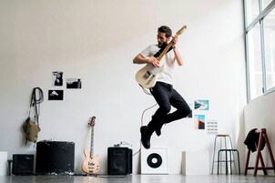Una persona tocando una guitarra eléctrica en una habitación con pared blanca, una ventana y diverso equipo musical.