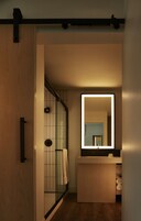 Un bagno moderno con porta in legno, cabina doccia in vetro, uno specchio grande e lavabo integrato.