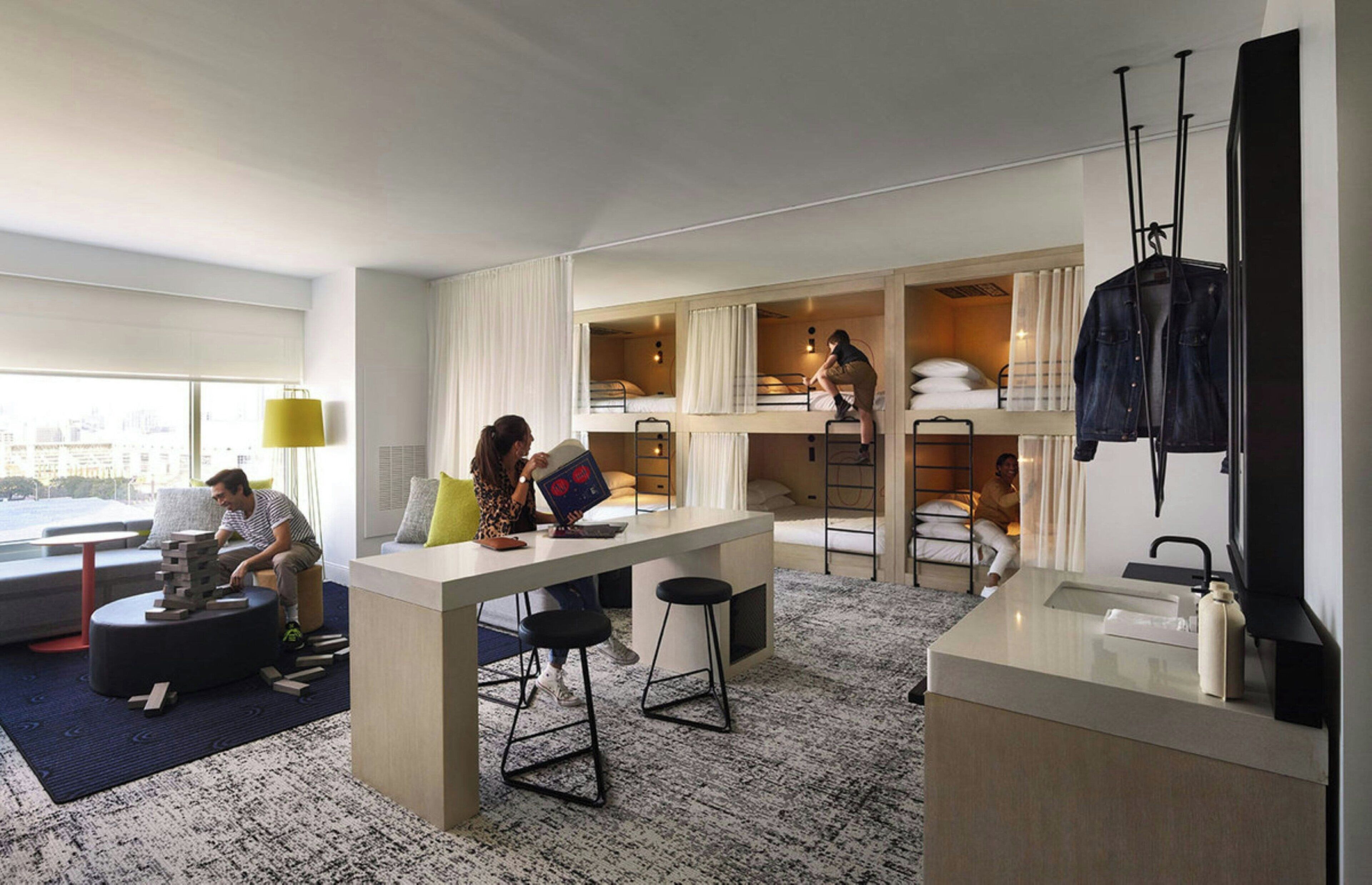 A modern hotel room with a bunk bed, a desk with a chair, a small seating area, and a large window.