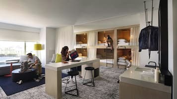 A modern hotel room with a bunk bed, a desk with a chair, a small seating area, and a large window.