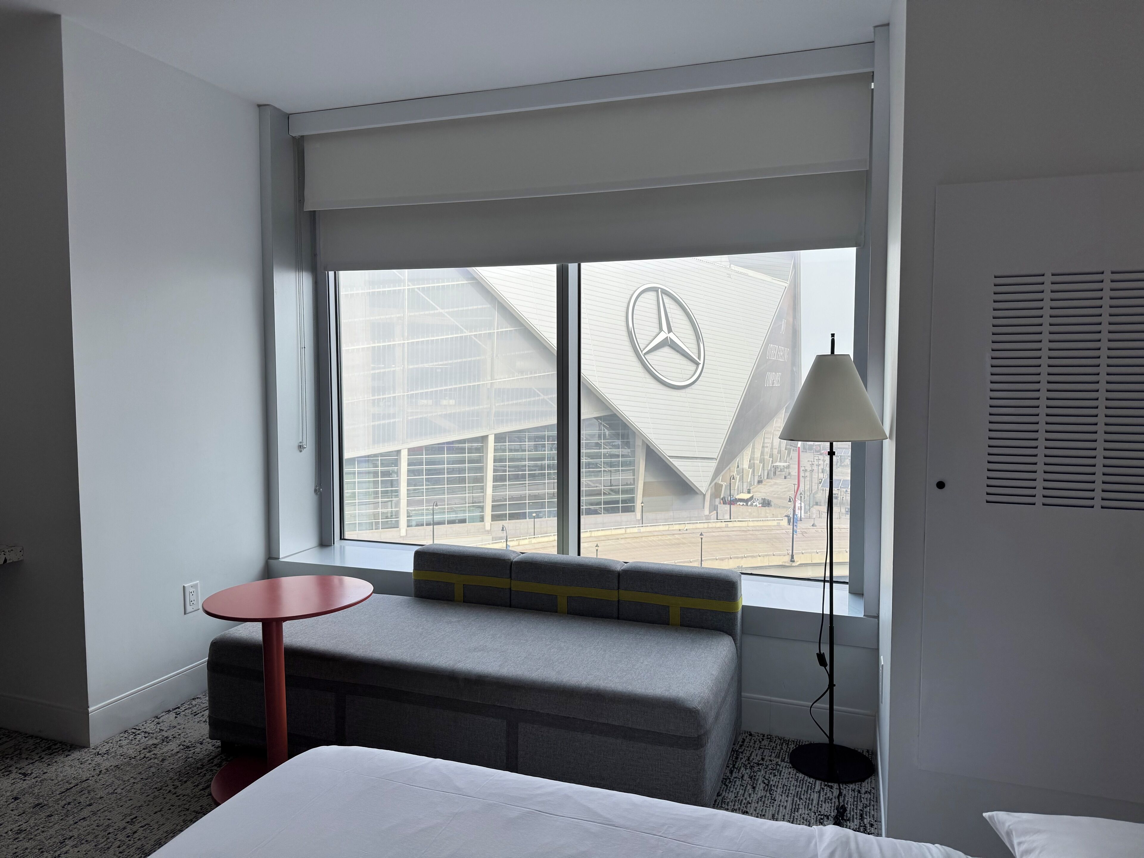 A hotel room with a large window, a bench, a small table, and a lamp.