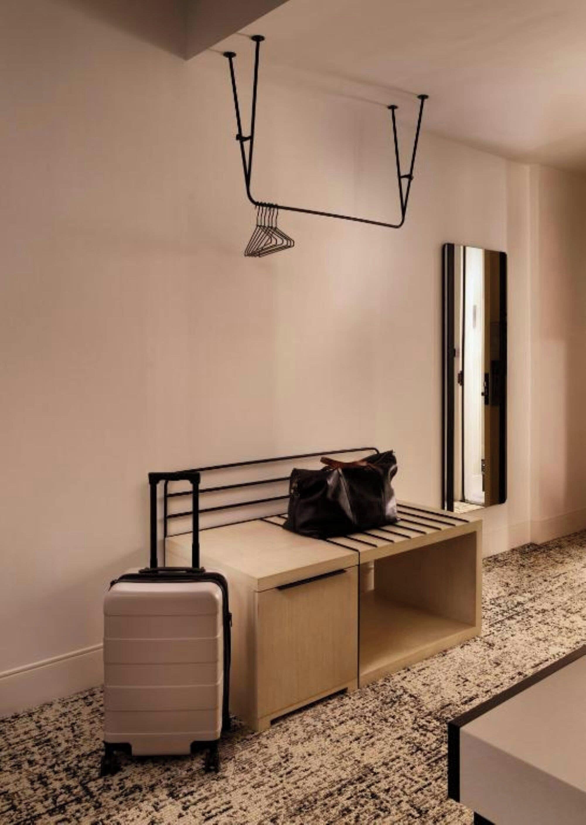 Une chambre d’hôtel avec un lit, une valise, un banc et un miroir.