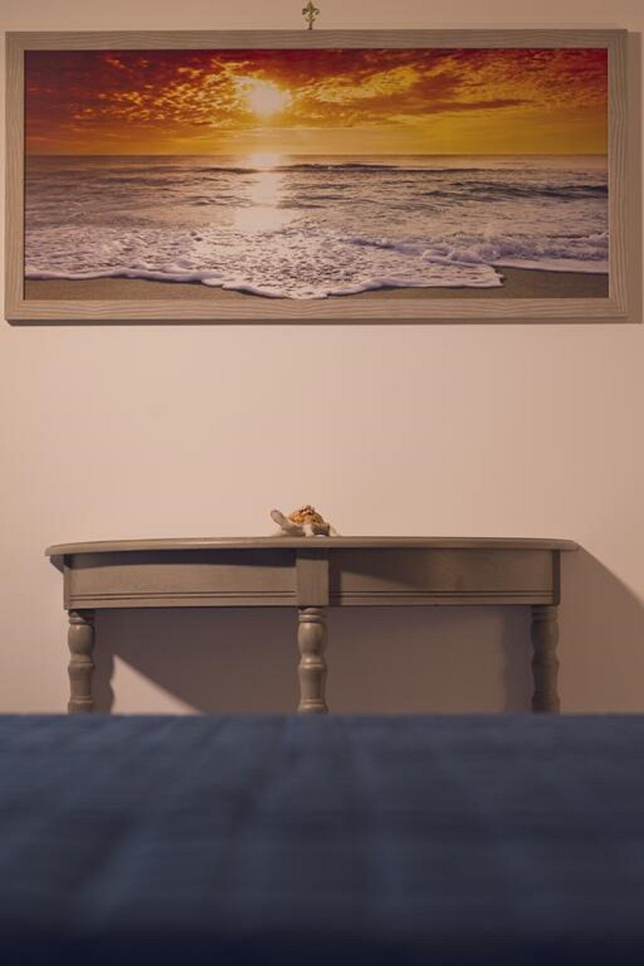 A room with a painting of a sunset over the ocean on the wall, a small wooden table with a cat on top, and a blue bedspread.