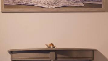 A room with a painting of a sunset over the ocean on the wall, a small wooden table with a cat on top, and a blue bedspread.