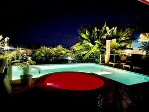 Vista nocturna de una piscina rodeada de plantas tropicales y un jacuzzi iluminado en rojo.