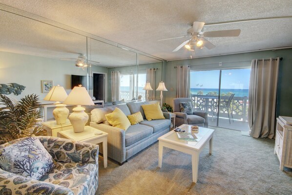 A living room with a sofa, coffee table, and chairs, a ceiling fan, and a view of the ocean through large windows. - El Matador 446 - FREE TICKETS!! Your stay includes free tickets to many area attractions! (Fort Walton Beach)
