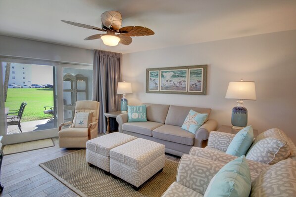 A living room with a sofa, armchairs, a coffee table, a ceiling fan, and a view of a grassy area through a sliding door.