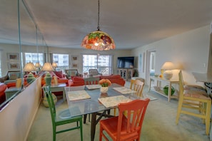 Un coin repas avec une table en verre, des chaises rouges et vertes, et un luminaire suspendu aux couleurs vives.