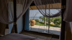A room with a bed, a large window with sheer curtains, and a view of a garden and palm trees. - Rushel Kivu Lodge (Boneza)
