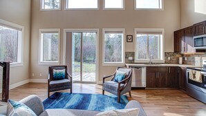 A spacious living room with a large window, a fireplace, and a kitchen area with dark wooden cabinets and a stone backsplash. - Modern Chalet Duplex - 2 (Mount Hood Village)