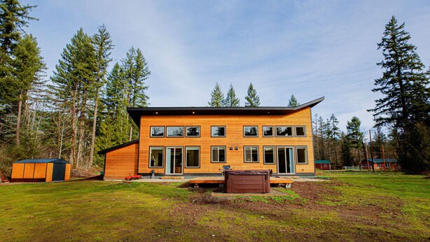 Een modern houten huis met twee verdiepingen en een ruime veranda, omgeven door een gazon en hoge bomen.