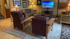 Uma sala de estar aconchegante com sofá, poltronas, mesa de centro e televisão. Há quadros nas paredes e um abajur.