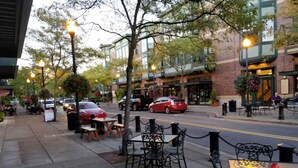 An outdoor cafe with tables and chairs, a street with cars, and buildings in the background. - Modern home in Orenco (Hillsboro)