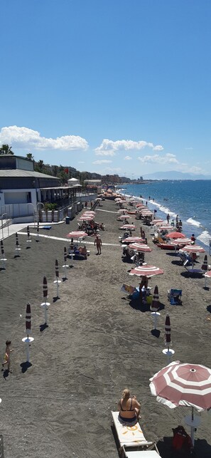 Una playa con hileras de sombrillas y sillas de descanso, un edificio con una bandera, y el mar al fondo.