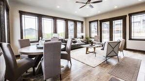 Uma sala de estar espaçosa com uma mesa de jantar redonda, cadeiras estofadas cinzas, um sofá e uma mesa de centro. Há grandes janelas que proporcionam luz natural e vista para o exterior.
