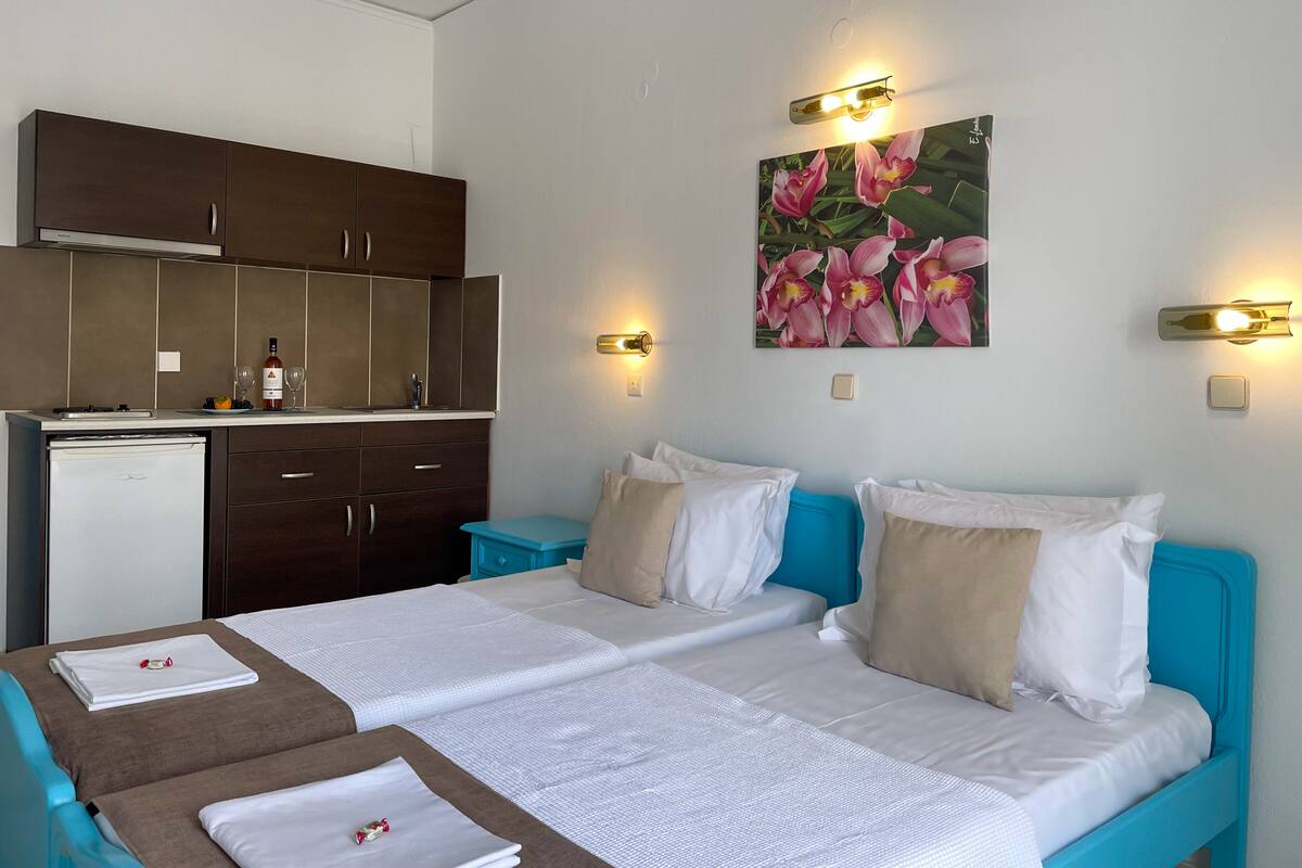 Quarto de hotel com cozinha compacta, duas camas com roupa de cama azul e bege, um quadro de flores cor-de-rosa e luminárias fixadas na parede.