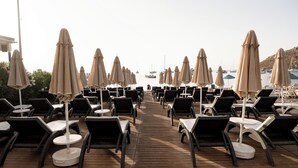 A wooden deck with rows of sun loungers and beige umbrellas, leading to a marina with boats. - Mare Garden Hotel Akyarlar (Bodrum)