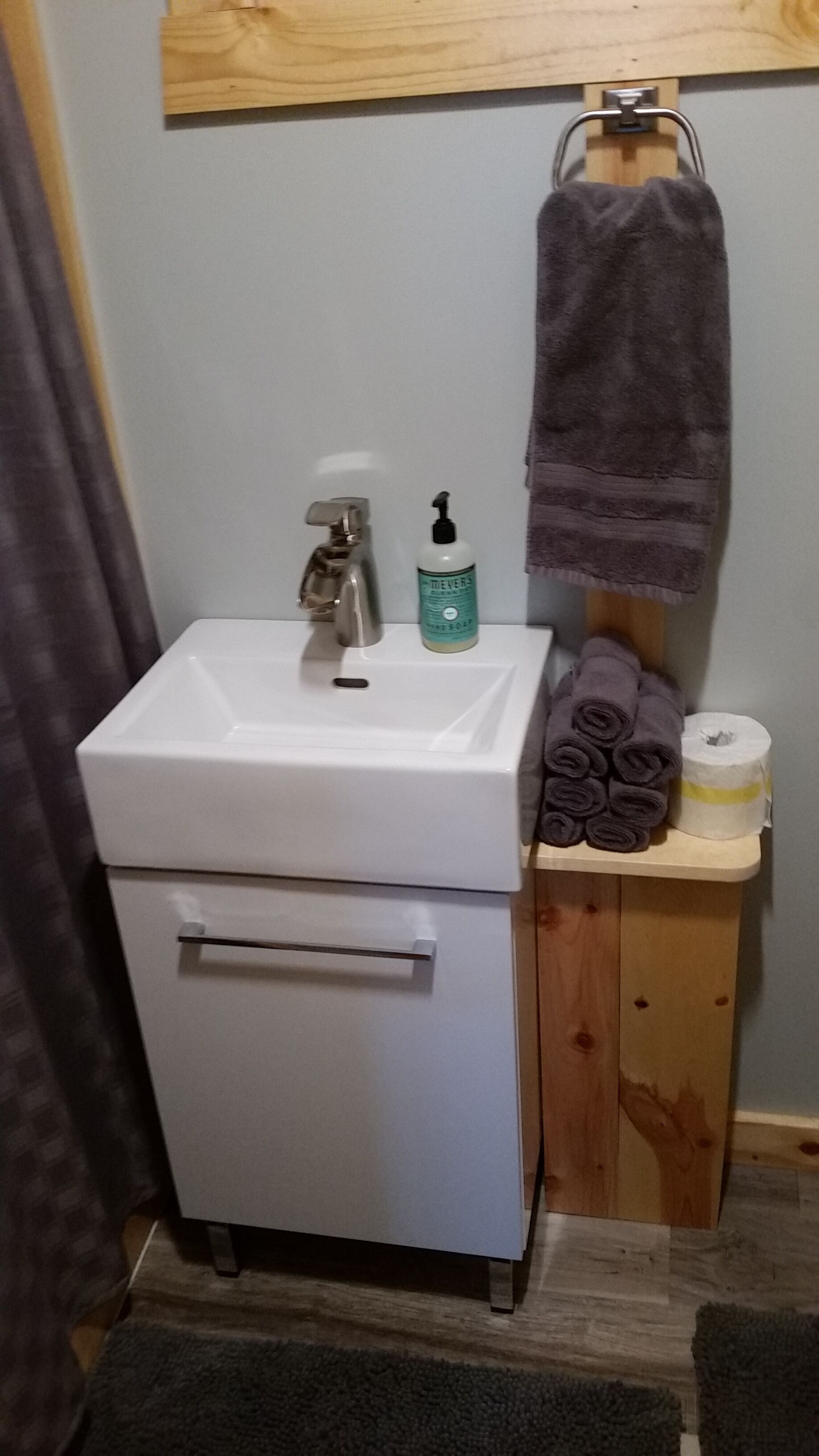 Un baño con un lavamanos blanco, un estante de madera con toallas y papel higiénico, y un perchero para toallas.