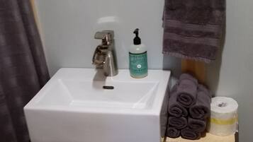 Un baño con un lavamanos blanco, un estante de madera con toallas y papel higiénico, y un perchero para toallas.