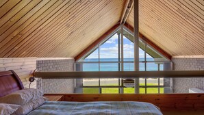 Quarto com teto de madeira, uma janela ampla com vista para a praia, cama com colcha xadrez e parede de tijolos.