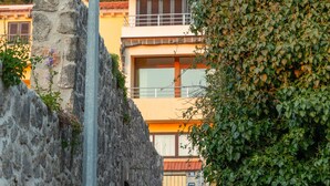 A stone wall with a metal lamp post, a staircase leading to a building with a red-tiled roof, and a lush green hedge.