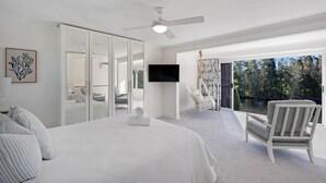 Un dormitorio con una cama grande, un ventilador de techo, un televisor, una silla y vistas a árboles.