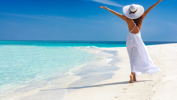 A person in a white dress and hat standing on a beach with arms outstretched, facing the ocean. - Calm Waters Waterfront Cottages @ Sussex Inlet, No. 2 Waterfront (Sussex Inlet)