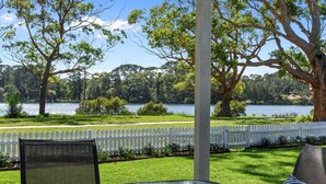 A glass table with outdoor seating, a white picket fence, and a view of a lake with trees. - Calm Waters Waterfront Cottages @ Sussex Inlet, No. 2 Waterfront (Sussex Inlet)