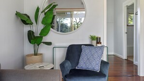 A room with a round mirror, a dark blue armchair with a patterned pillow, a large potted plant, and a small round table with a white marble top. - Calm Waters Waterfront Cottages @ Sussex Inlet, No. 2 Waterfront (Sussex Inlet)