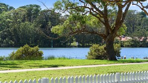 A park with a lake, a white picket fence, and trees. - Calm Waters Waterfront Cottages @ Sussex Inlet, No. 2 Waterfront (Sussex Inlet)