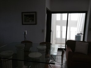 Une pièce faiblement éclairée, avec une table à manger en verre, des chaises, un canapé et une grande fenêtre munie de stores.