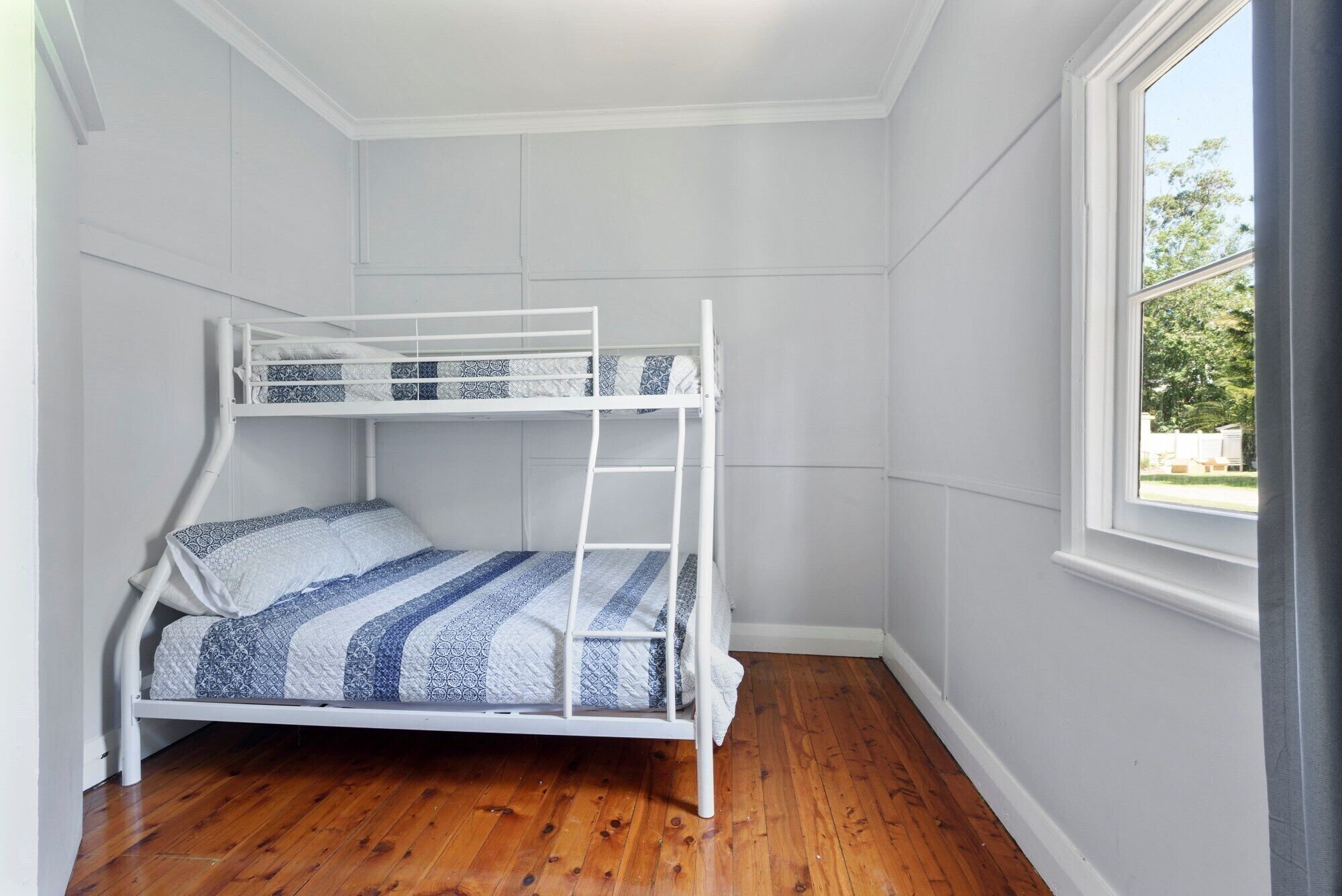 Una cama litera con estructura blanca y ropa de cama estampada en una habitación con suelo de madera y una ventana.