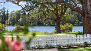 Una vista serena de un lago, con cerca blanca de piquetes, vegetación exuberante y un gran árbol.