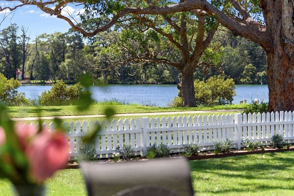 A serene lakeside view with a white picket fence, lush greenery, and a large tree.