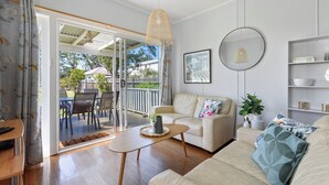 A living room with a sofa, a coffee table, and a dining area with chairs and a table. There is a sliding glass door leading to an outdoor patio. - Calm Waters Waterfront Cottages @ Sussex Inlet, No. 5 Garden View (Sussex Inlet)