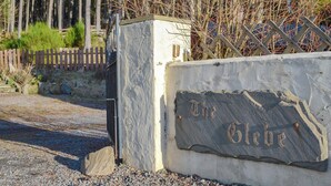 Un letrero de piedra con la inscripción “The Gibe” colocado en una pared de concreto, rodeado de una cerca de madera y árboles.