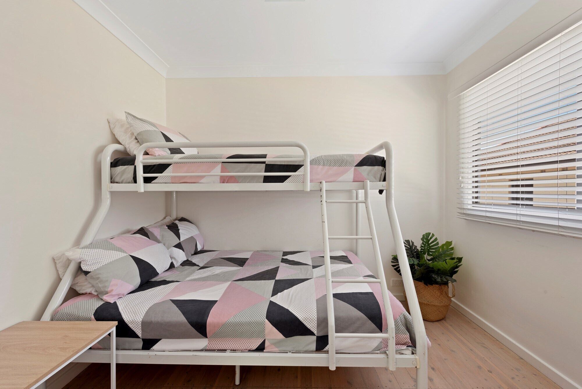 A bunk bed with geometric patterned bedding, a small table, and a potted plant in a corner room with a window and blinds.