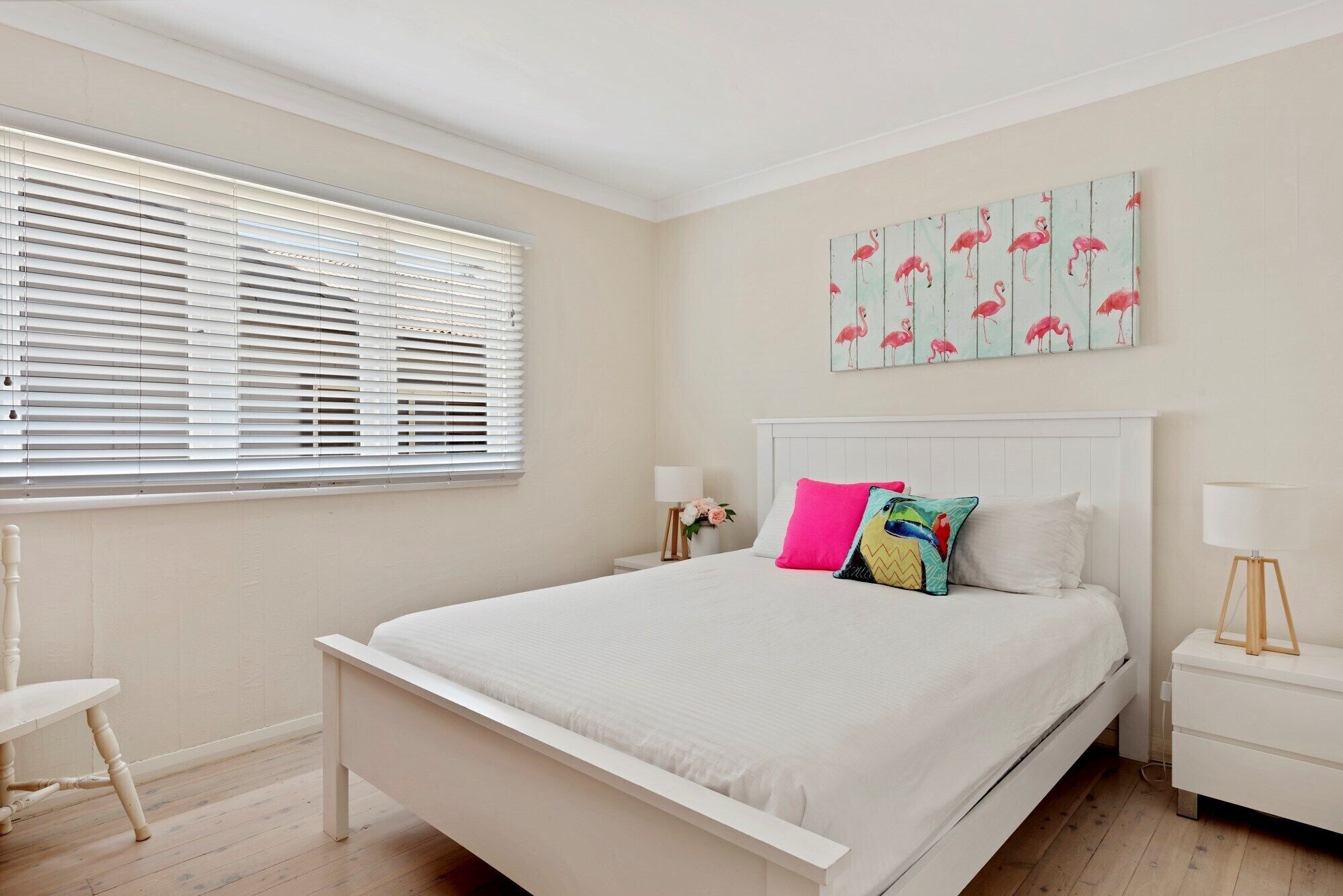 Quarto com cama branca, mesa de cabeceira com abajur, cadeira e janela com persianas.