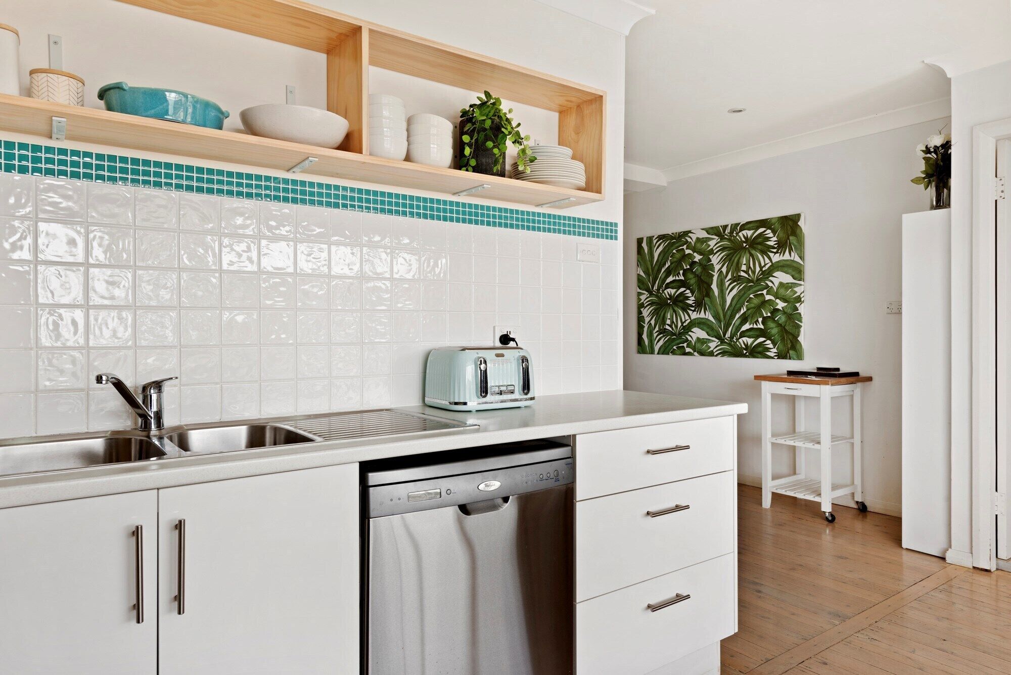 A modern kitchen with white cabinets, a stainless steel sink, a dishwasher, and a wooden shelf with decorative items.