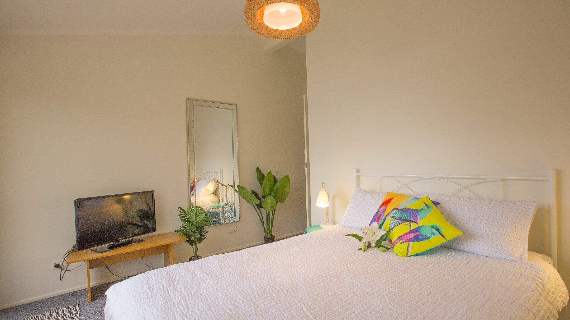 Et soveværelse med en seng, et fjernsyn på et træstel, et spejl, en potteplante og en sengetårnlampe.