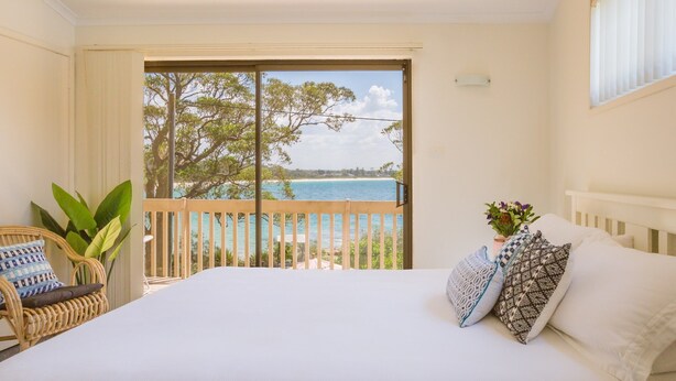 Un dormitorio con una cama grande, vistas al mar, un balcón con una silla de mimbre y una planta en maceta.