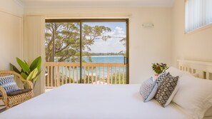 A bedroom with a large bed, a view of the sea, a balcony with a wicker chair, and a potted plant. - Driftwood - Beachfront location, pet friendly, families (Vincentia)