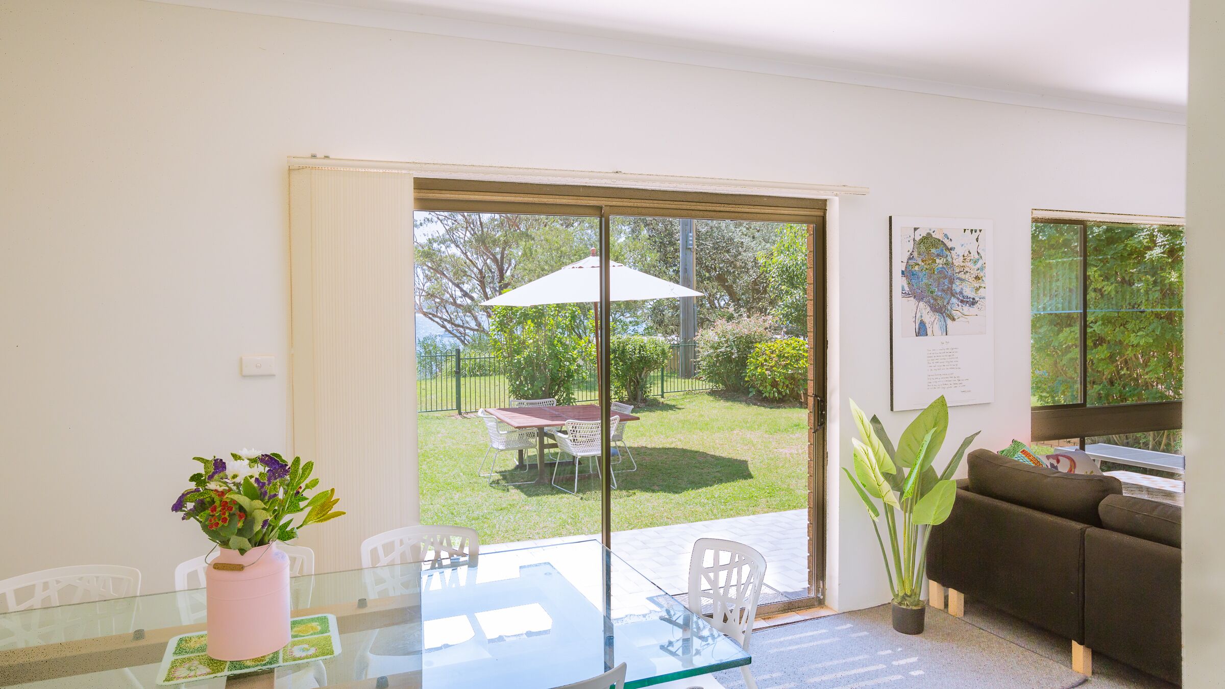 Un salon moderne avec une table en verre, des chaises blanches, un canapé marron et une porte-fenêtre coulissante donnant sur un jardin.