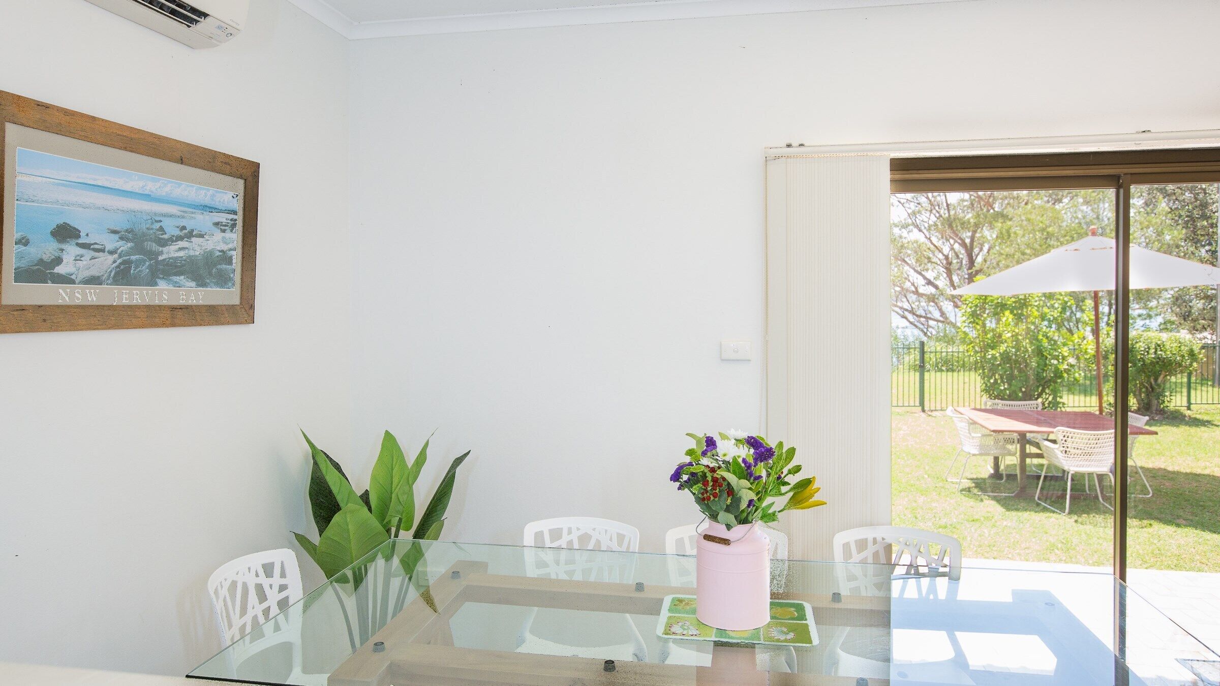 A dining area with a glass table, chairs, a pink vase with flowers, and a framed picture on the wall.