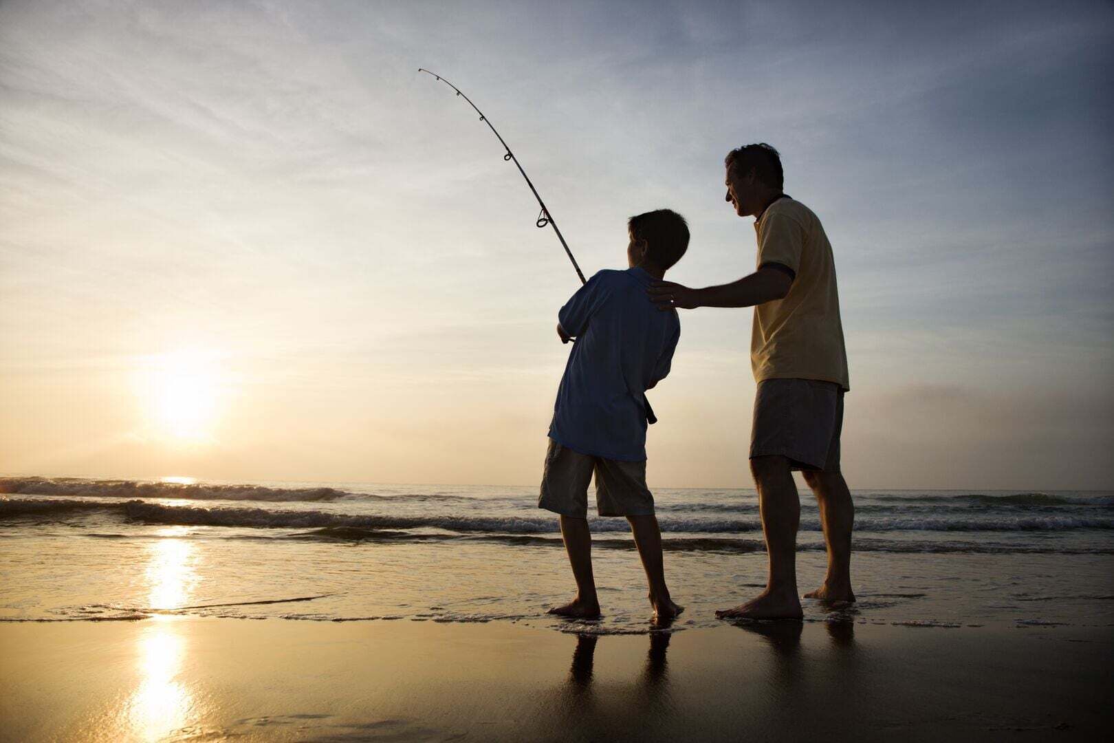 Two people fishing on a beach with the sun setting in the background.