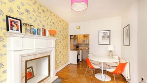 A room with a white fireplace, a dining table with orange chairs, a kitchen in the background, and a patterned wallpaper. - Stunning Apartment in Royal Mile (Edinburgh)