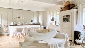 A cozy, well-lit room with a white sofa, a dining table with chairs, a kitchen area, and a large window with curtains. - Charming fisherman's house near the beach and the village center (Veules-les-Roses)